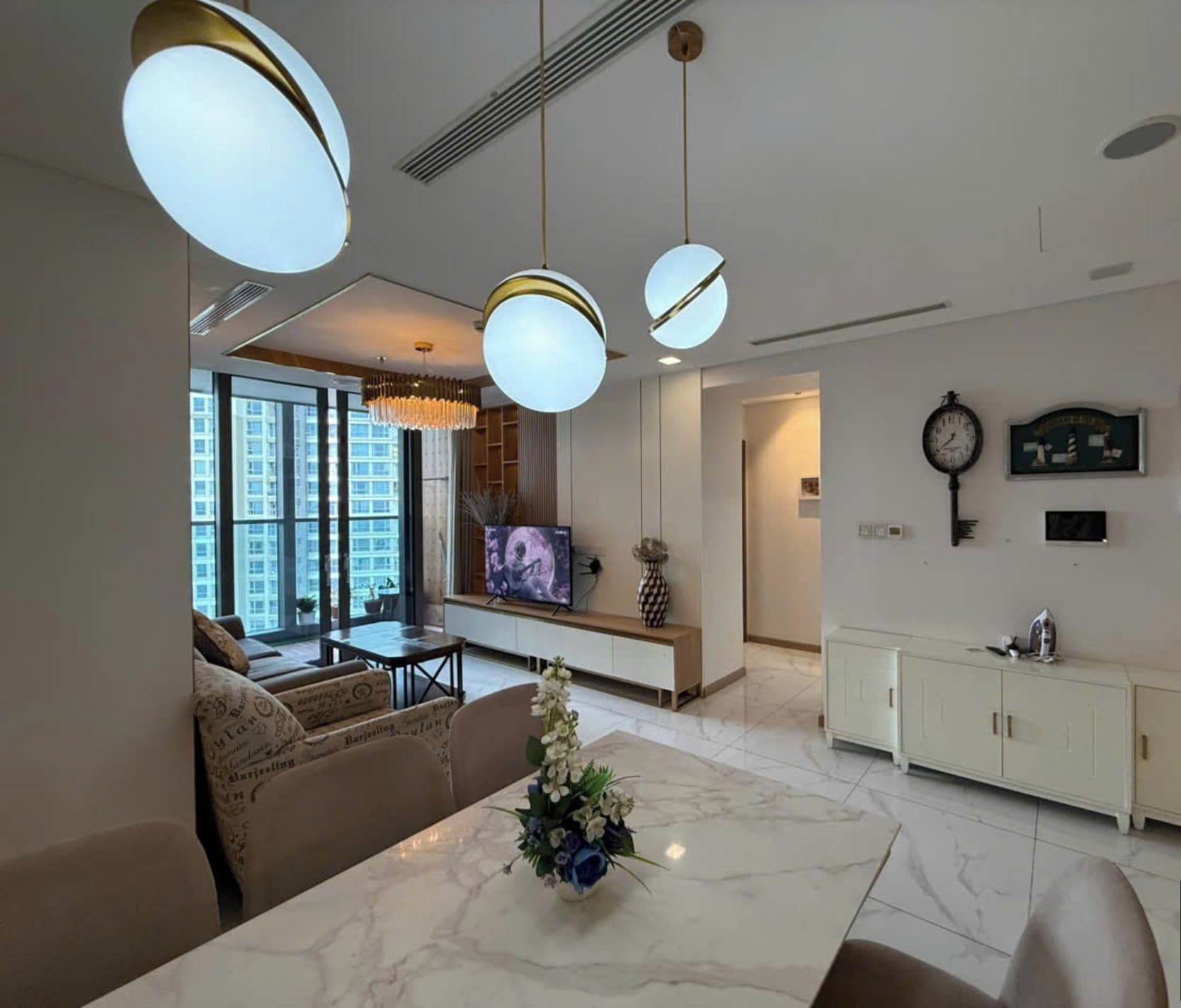 Ein modernes Wohnzimmer mit einem Marmortisch, einer Couch und einem Fernseher. Es gibt Pendelleuchten, eine Uhr und gerahmte Bilder an der Wand.