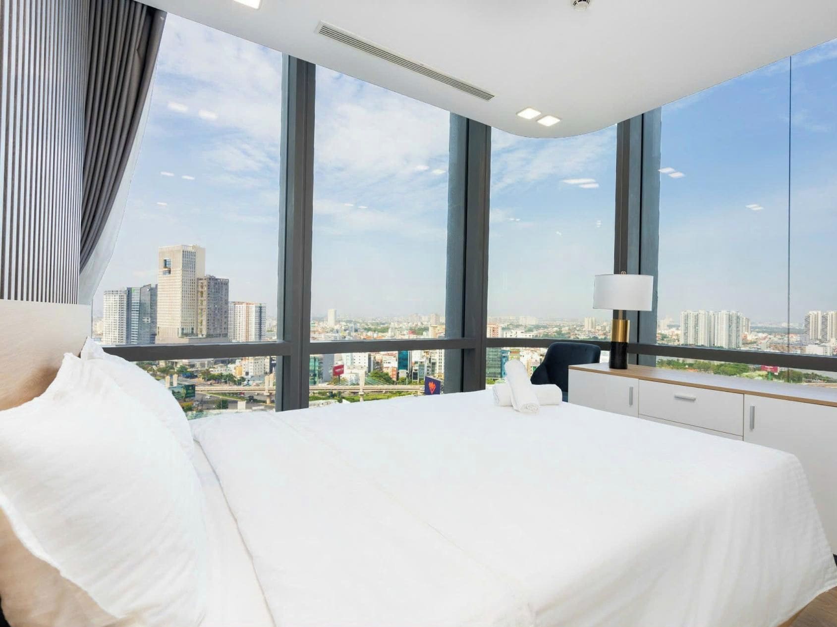 A modern hotel room with a large bed, a nightstand, and a view of a cityscape through floor-to-ceiling windows.