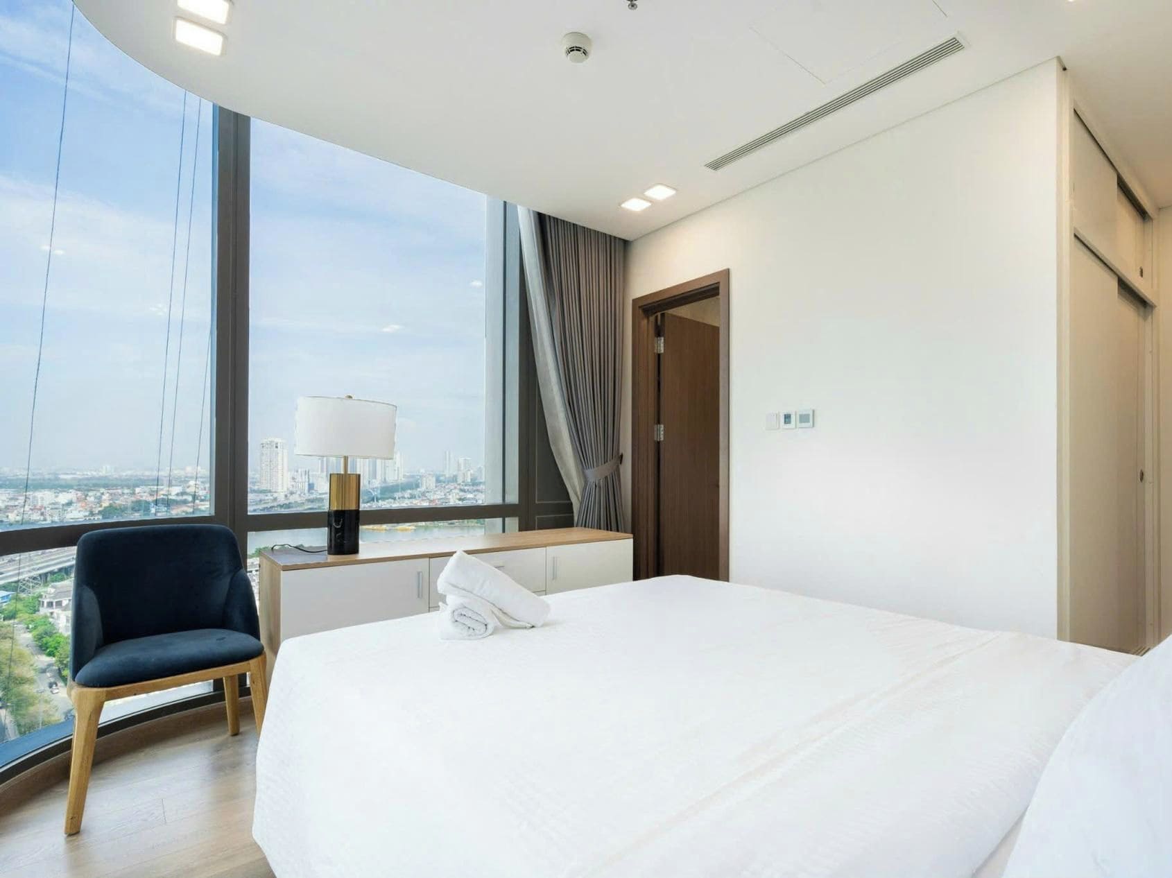 Camera d'albergo con un letto grande, una scrivania con una lampada, una sedia e vista sulla città attraverso ampie finestre.