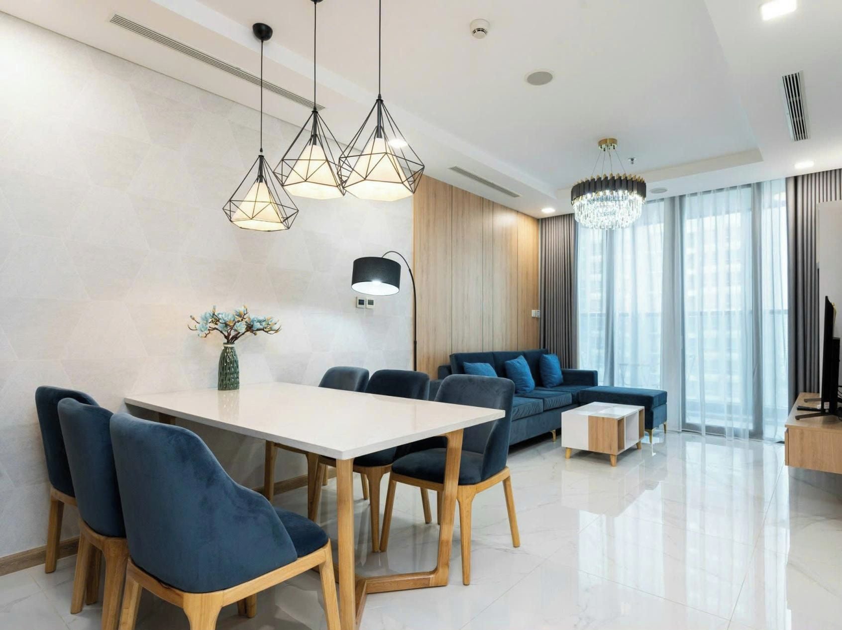 A modern living room with a dining area, featuring a large table, blue sofas, and a chandelier.