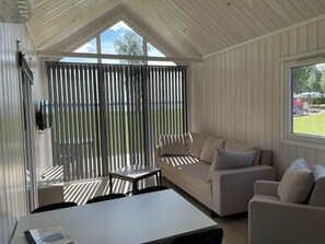 A modern room with a high ceiling, a large window, a sofa, a table, and a TV mounted on the wall. - Gökaskratts Camping (Hovmantorp)