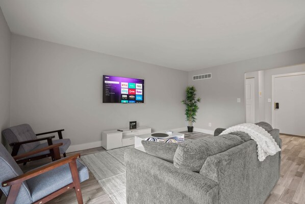 Un salón moderno con un sofá gris, una silla de madera, una mesita auxiliar blanca y un televisor de pantalla plana montado en la pared mostrando servicios de streaming.