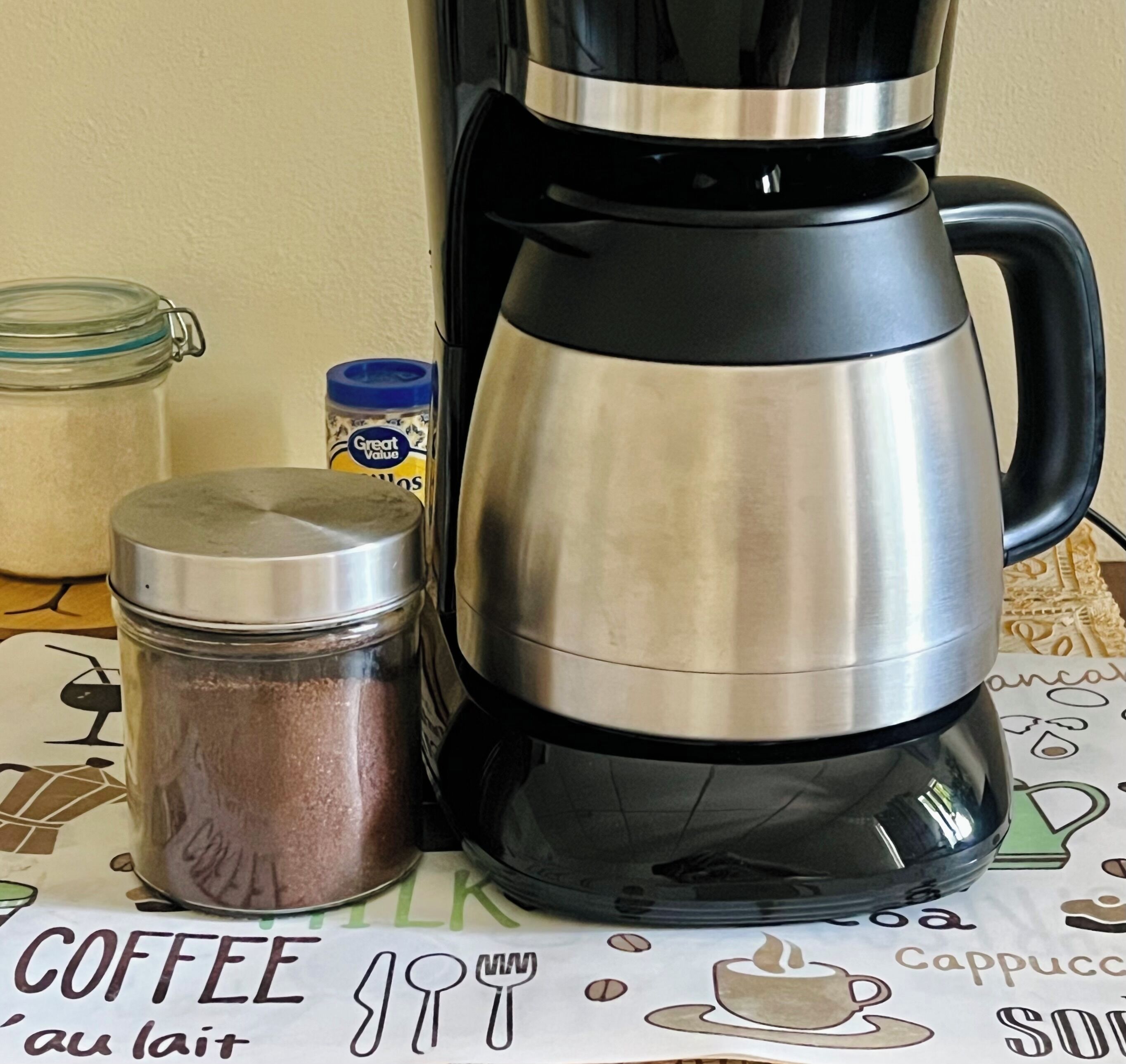 A coffee maker, a jar of coffee, and a container of creamer on a table with a coffee-themed placemat.