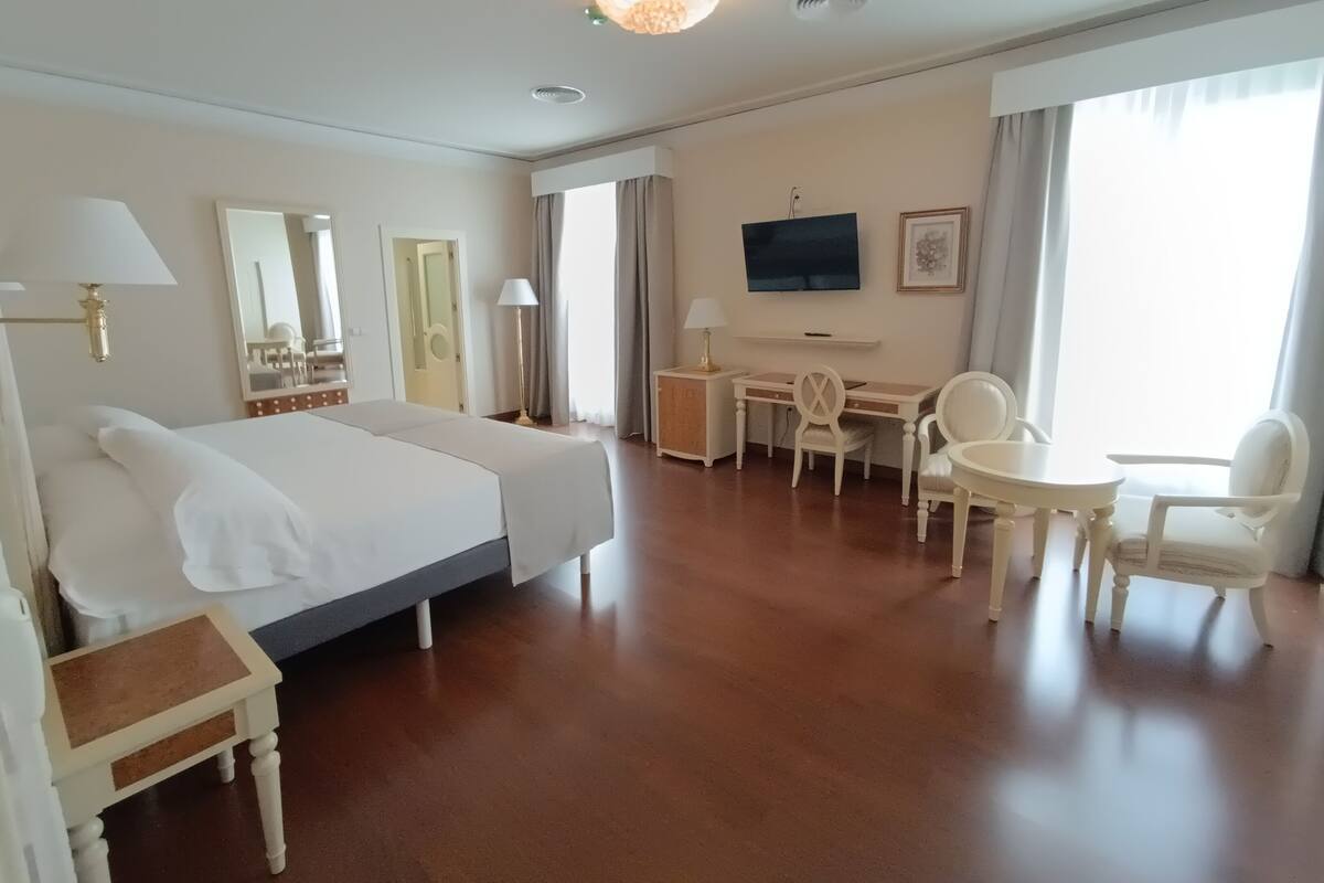 Hotelkamer met groot bed, bureau, stoel, klein tafeltje, televisie en raam met gordijnen.