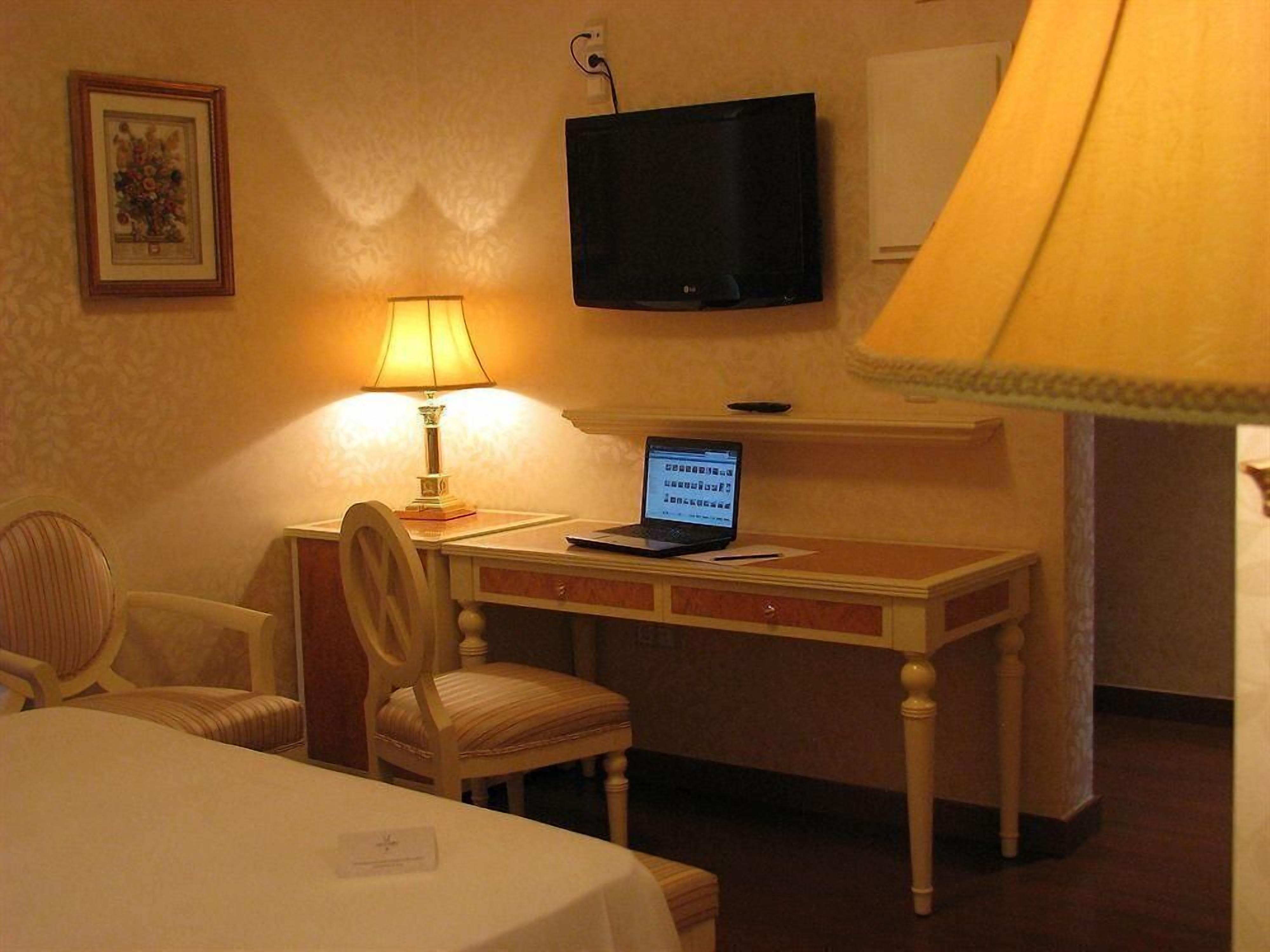Une chambre d’hôtel avec un lit, un bureau avec un ordinateur portable, une chaise, une lampe, une télévision et un tableau encadré au mur.