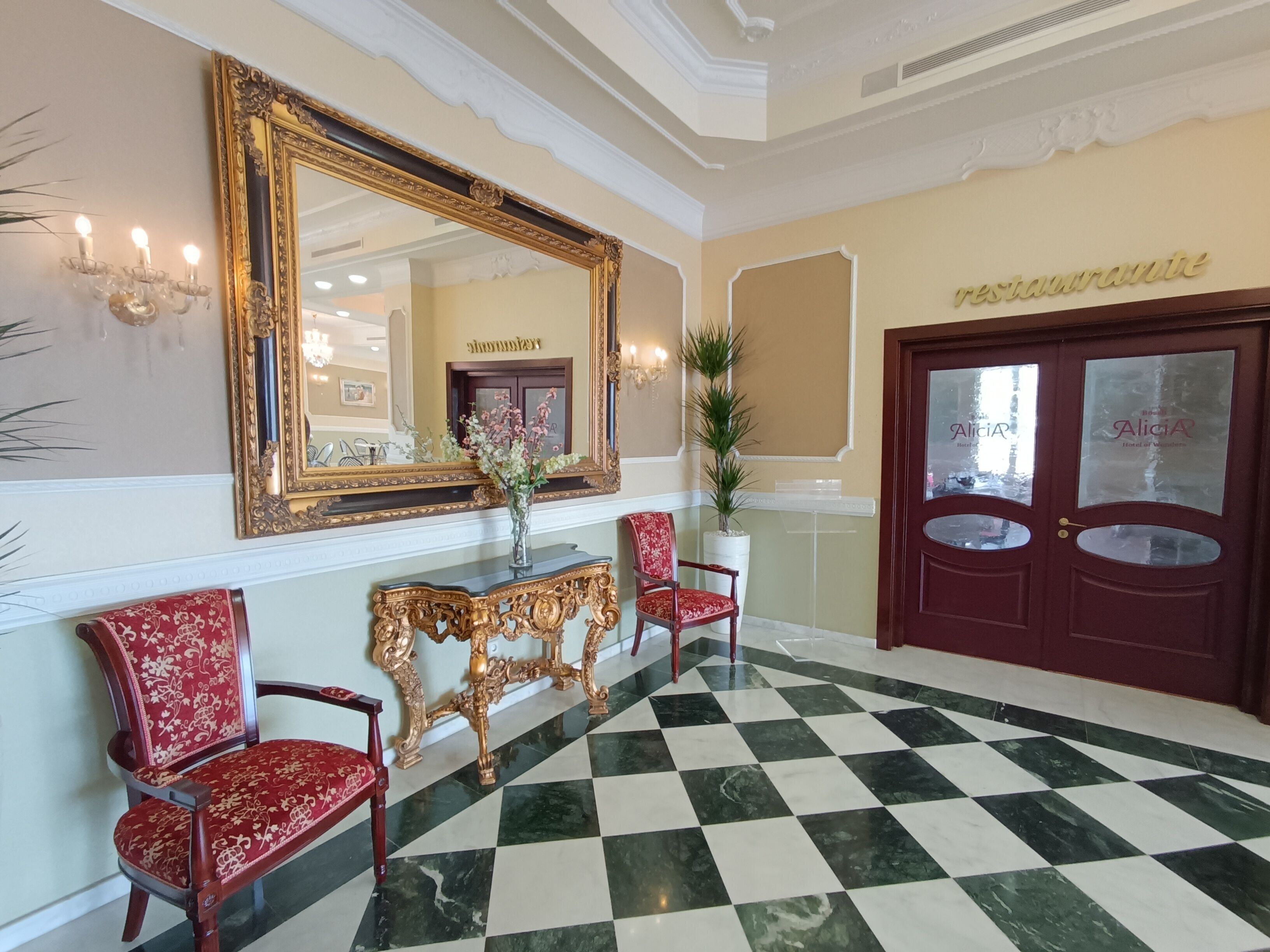 A lobby with a checkered floor, a large ornate mirror, red upholstered chairs, a console table, and a door with signage.