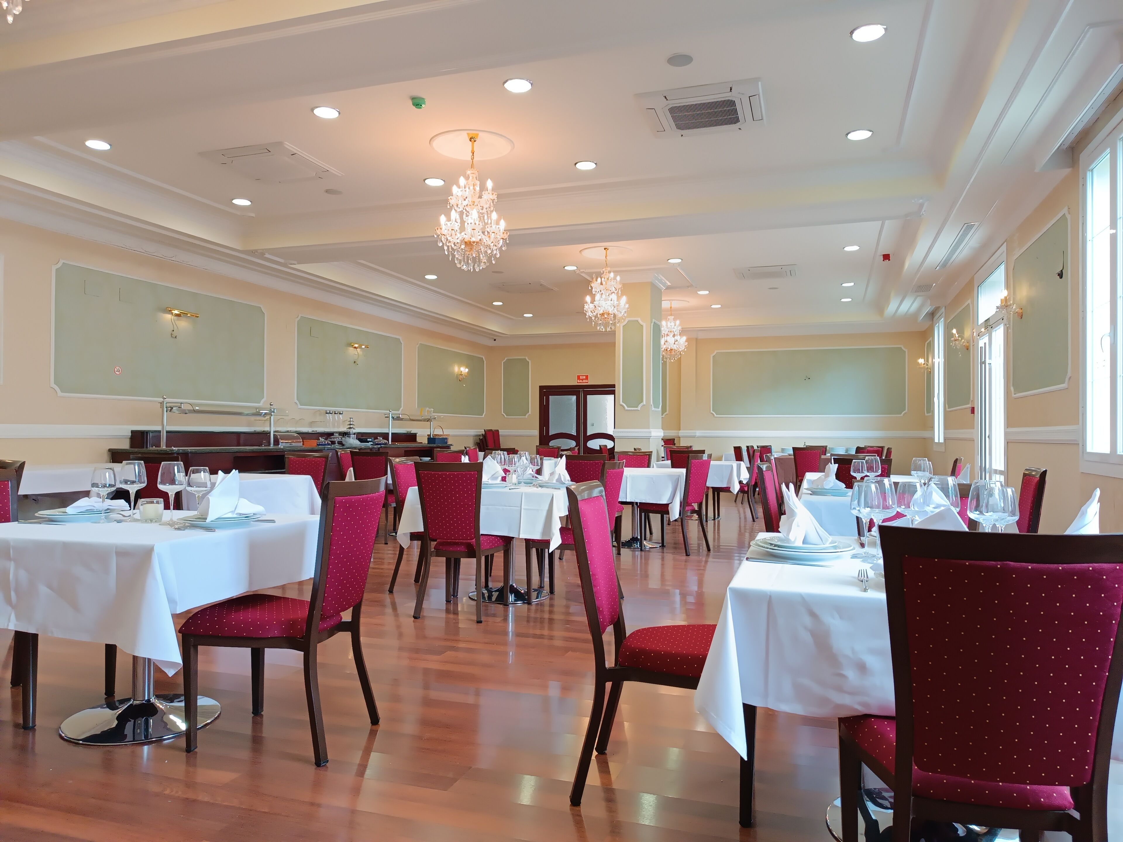 A spacious dining area with multiple tables and chairs, chandeliers, and large windows.