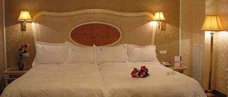 A large bed with a headboard, two bedside lamps, a bedside table with a vase of flowers, and a bench at the foot of the bed.