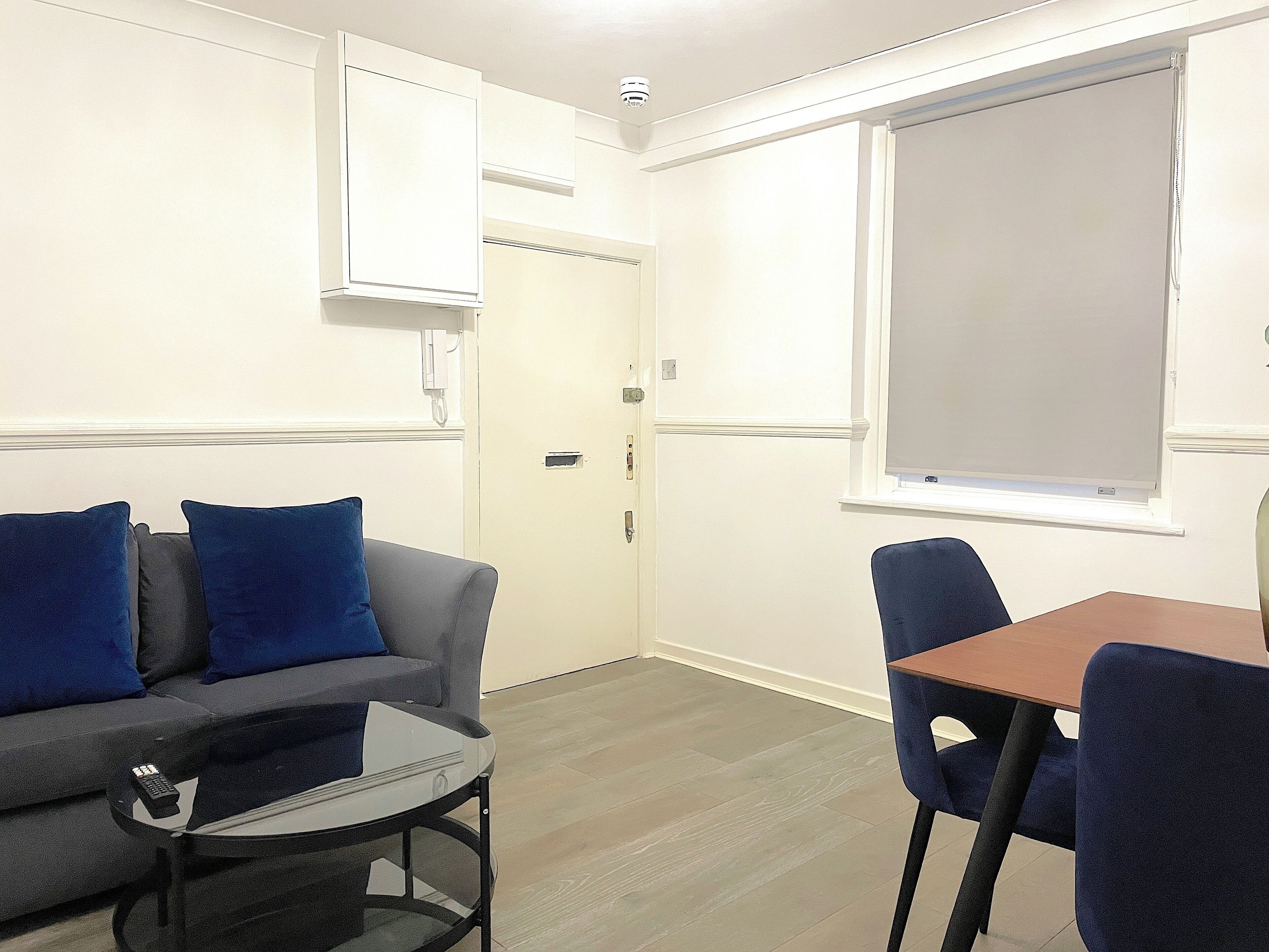 Une pièce avec un canapé gris, des coussins bleus, une petite table, une chaise bleue, une fenêtre avec un store et une porte avec une serrure.