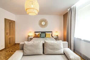 Ein Schlafzimmer mit einem Bett, zwei Nachttischen mit Lampen, einer Holztür, einem runden Spiegel an der Wand und einem Fenster mit Vorhängen.