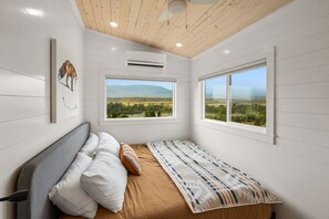 A bedroom with a bed, pillows, a patterned blanket, a large window with a view of a landscape, and an air conditioning unit. - One More Cast Cabin -a short walk to the Yellowstone River! (Pray)
