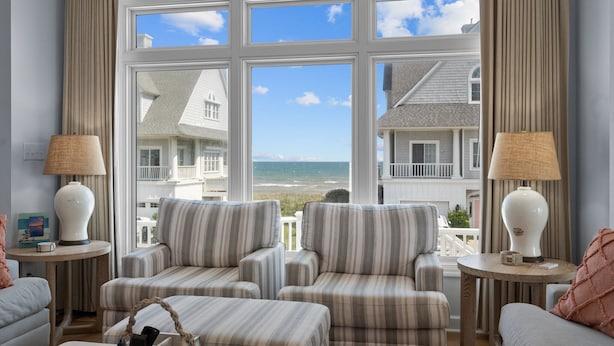 A room with two striped armchairs, a sofa, and a view of the ocean through large windows. - Our Happy Place #170 Ocean & Sound Views, Hot Tub, Pool & Beach Access (North Topsail Beach)
