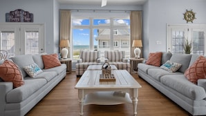 A spacious living room with two grey sofas, a white coffee table, and a view of a house through large windows. - Our Happy Place #170 Ocean & Sound Views, Hot Tub, Pool & Beach Access (North Topsail Beach)