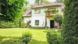Ein zweistöckiges Haus mit grüner Fassade, einem Balkon mit Holzgeländer und einem gepflegten Garten mit Rasen und verschiedenen Pflanzen.
