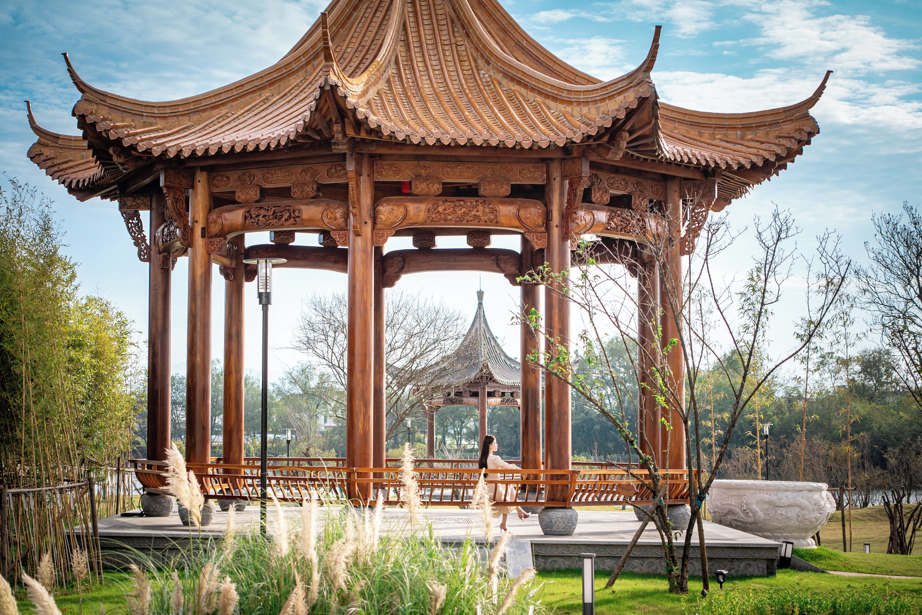 A traditional pavilion with ornate wooden pillars and a tiled roof, surrounded by greenery and a body of water.