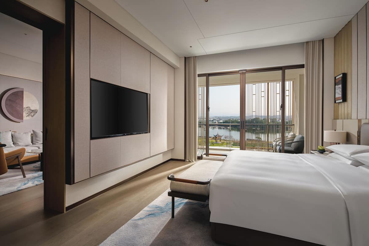 Une chambre d’hôtel moderne dotée d’un grand lit, d’un téléviseur à écran plat fixé au mur, d’un coin salon avec un canapé et un fauteuil, et d’un balcon offrant une vue sur la ville.