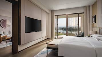 A modern hotel room with a large bed, a flat-screen TV mounted on the wall, a seating area with a sofa and armchair, and a balcony with a view of the city.