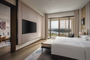 Ein modernes Hotelzimmer mit einem großen Bett, einem flachbildfernseher an der Wand, einer Sitzecke mit Sofa und Sessel sowie einem Balkon mit Blick auf die Stadt.