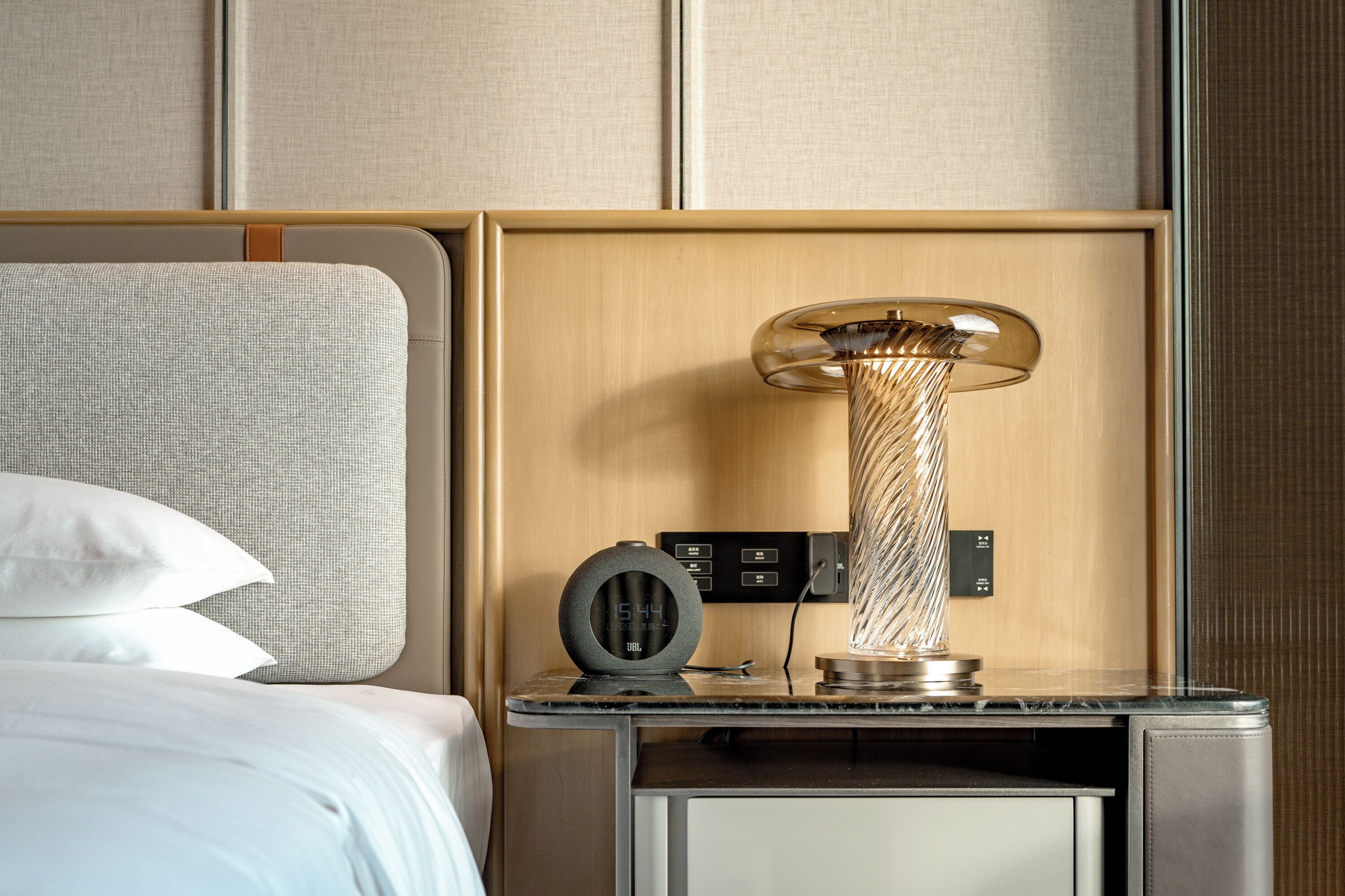 A hotel room with a bed, a bedside table, a lamp, and a control panel.