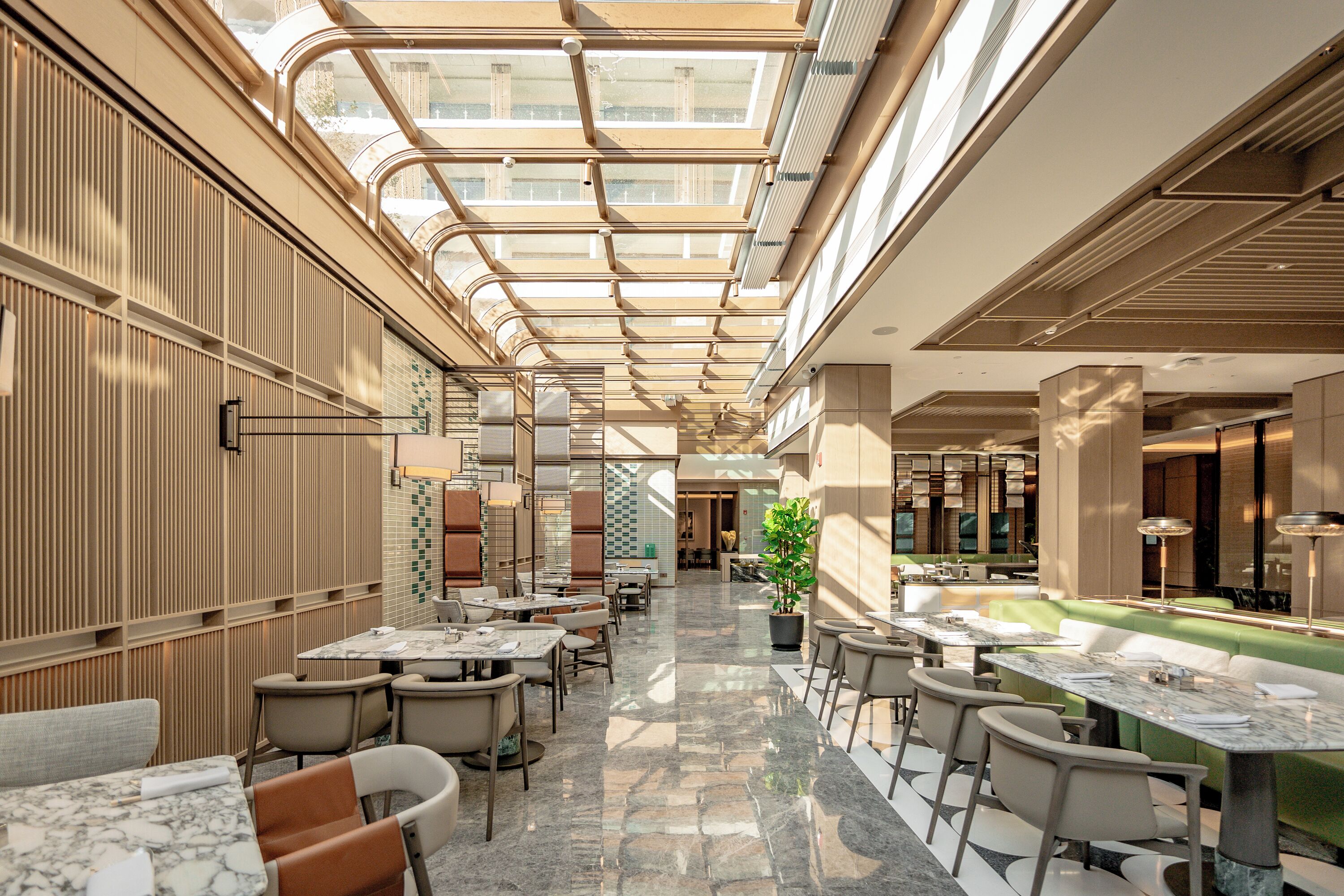 A modern indoor space with a central open area, surrounded by tables and chairs, and a glass ceiling allowing natural light.