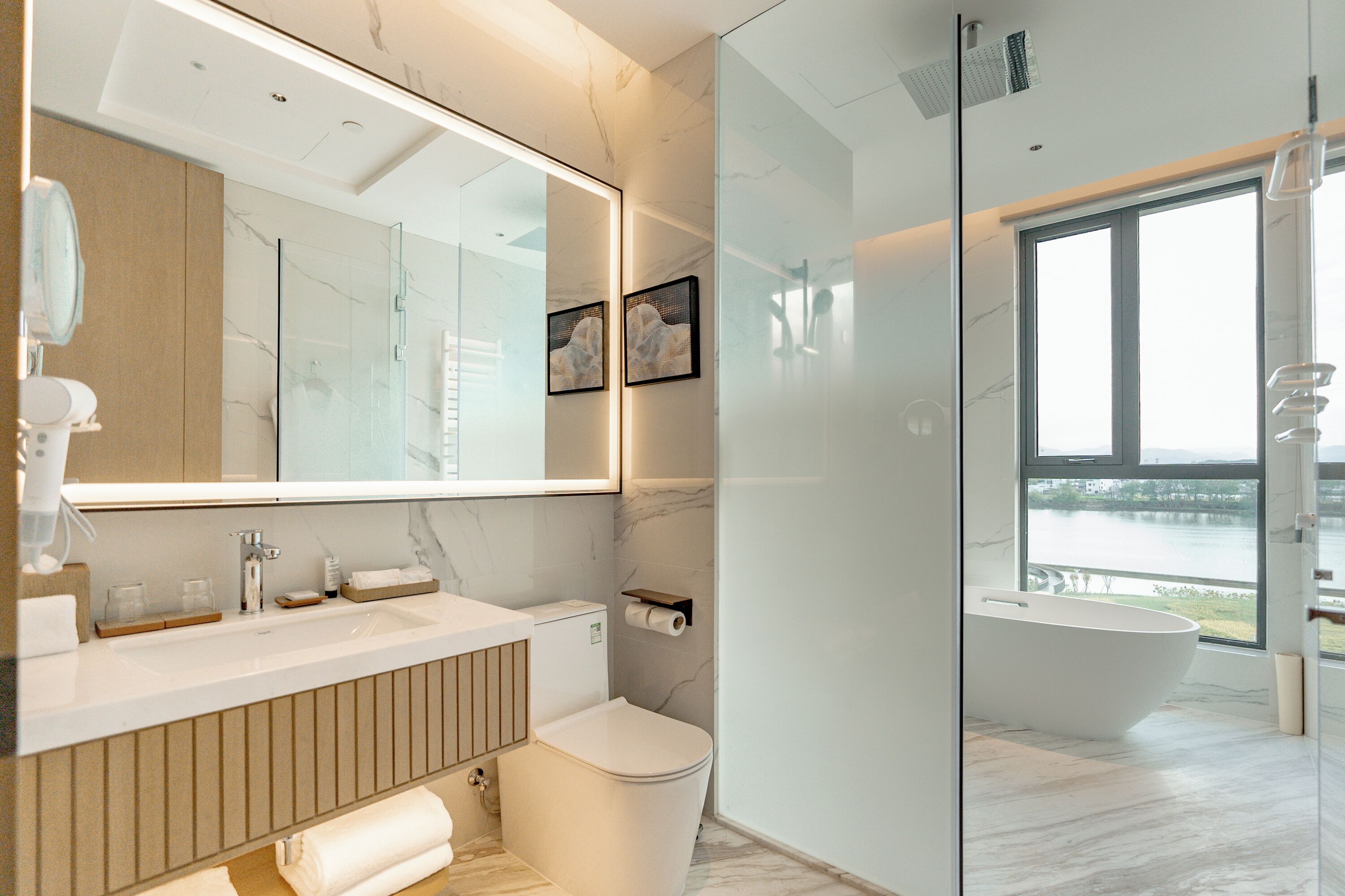 A modern bathroom with a glass-enclosed shower, a freestanding bathtub, a large mirror, and a view of a body of water.