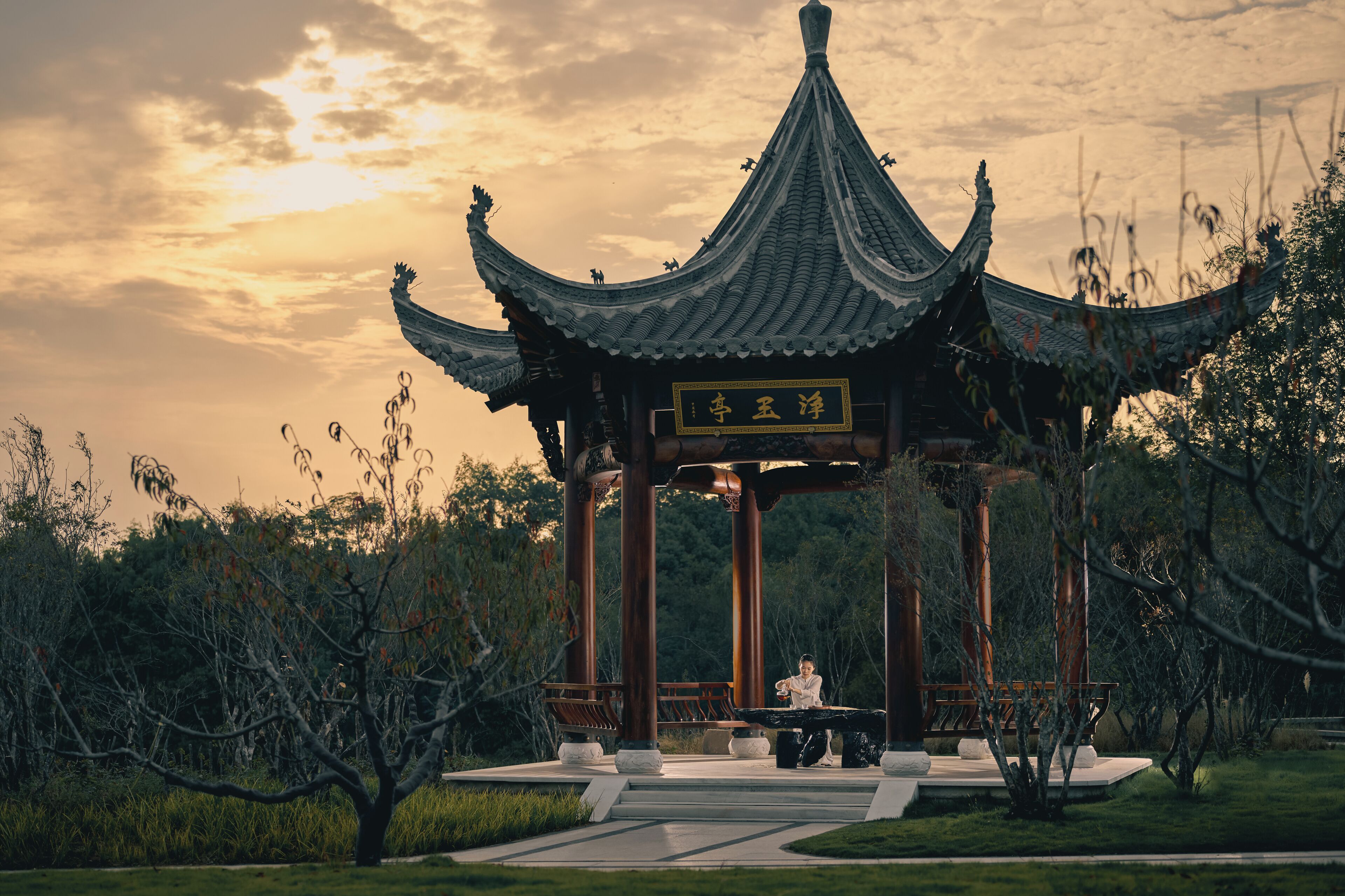 A traditional pavilion with a curved roof, surrounded by greenery and a pathway.