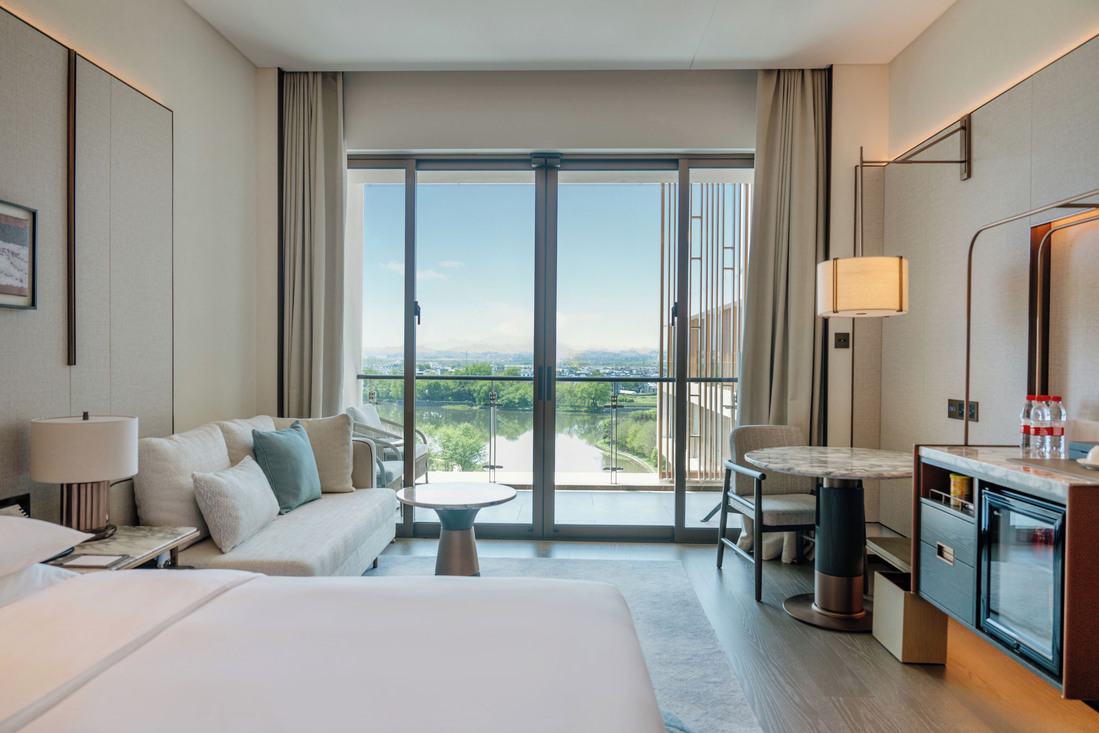 Une chambre d’hôtel comprenant un lit, un canapé, une petite table, un bureau et une vue sur le paysage urbain, visible à travers de grandes portes vitrées.