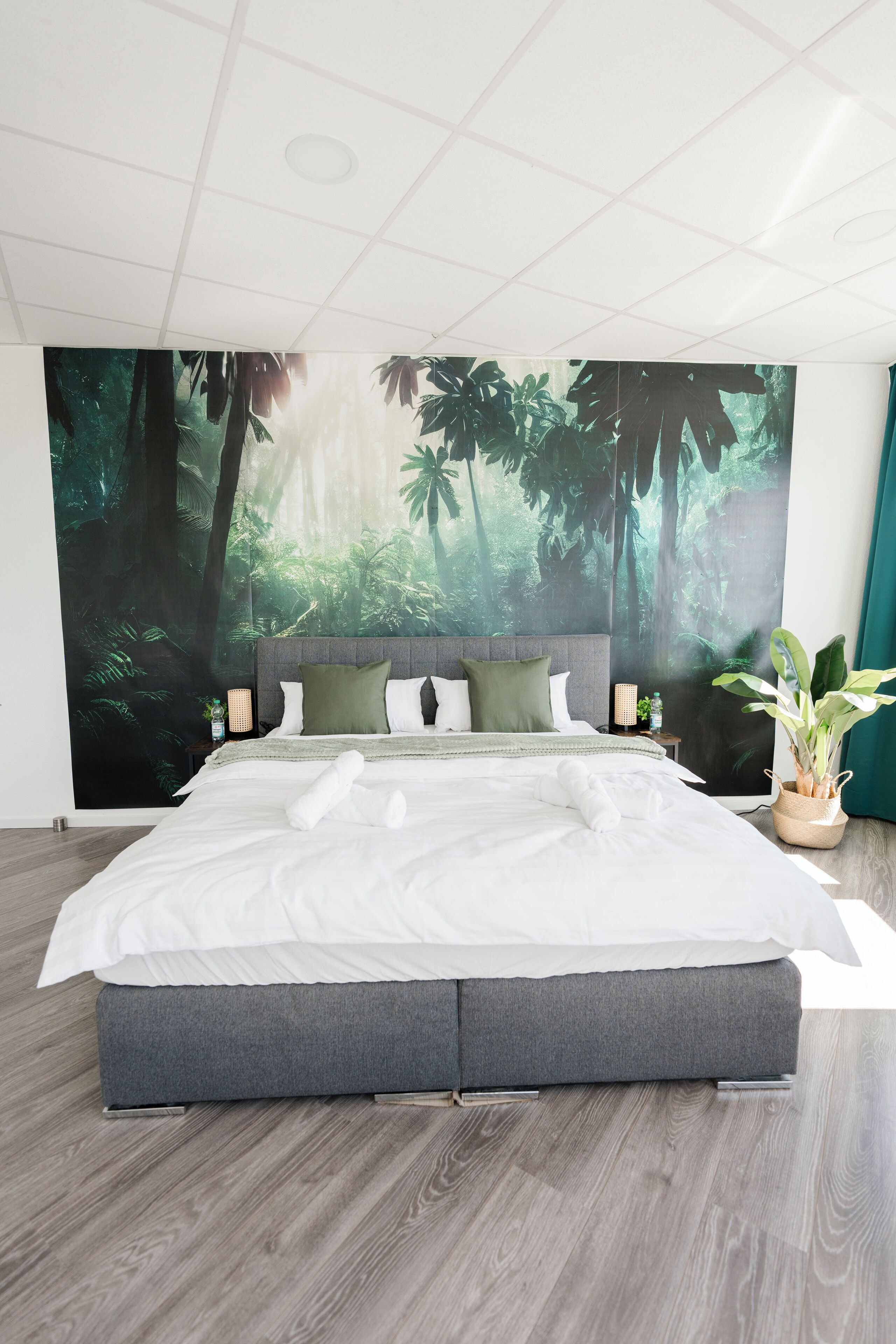 Une chambre à coucher moderne avec un grand lit, une fresque murale sur le thème de la forêt, une table de chevet avec une lampe et une plante en pot.