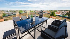Un patio al aire libre con una mesa de vidrio, sillas de mimbre negro y una parrilla.