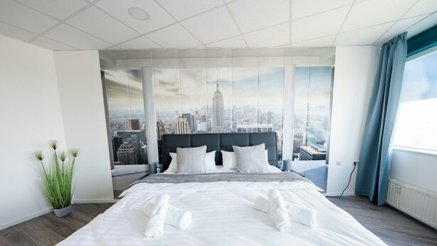 Habitación de hotel moderna con una cama grande, vista panorámica de la ciudad a través de una ventana y una planta en maceta.