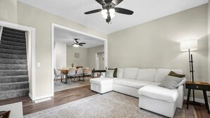 Una sala de estar con un sofá modular, una zona de comedor con una mesa y sillas, una escalera y un ventilador de techo.