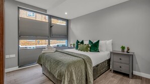 A modern bedroom with a large window, a bed with green bedding, a nightstand, and a small plant.