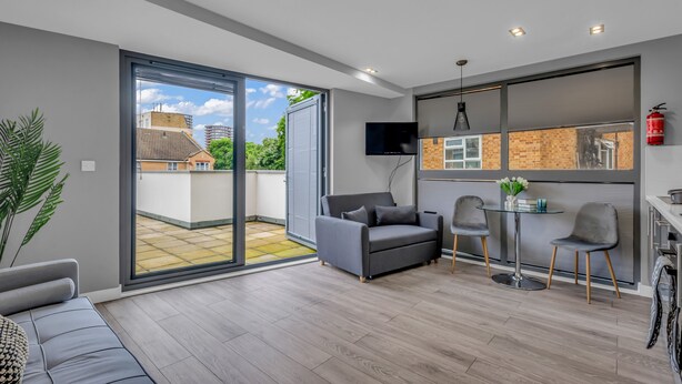A modern living room with a grey sofa, a small table with two chairs, a TV mounted on the wall, and a sliding glass door leading to a balcony.