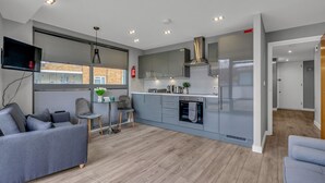 A modern kitchen with grey cabinets, a built-in oven, and a stainless steel range hood. There is a dining area with a grey sofa and a small table.