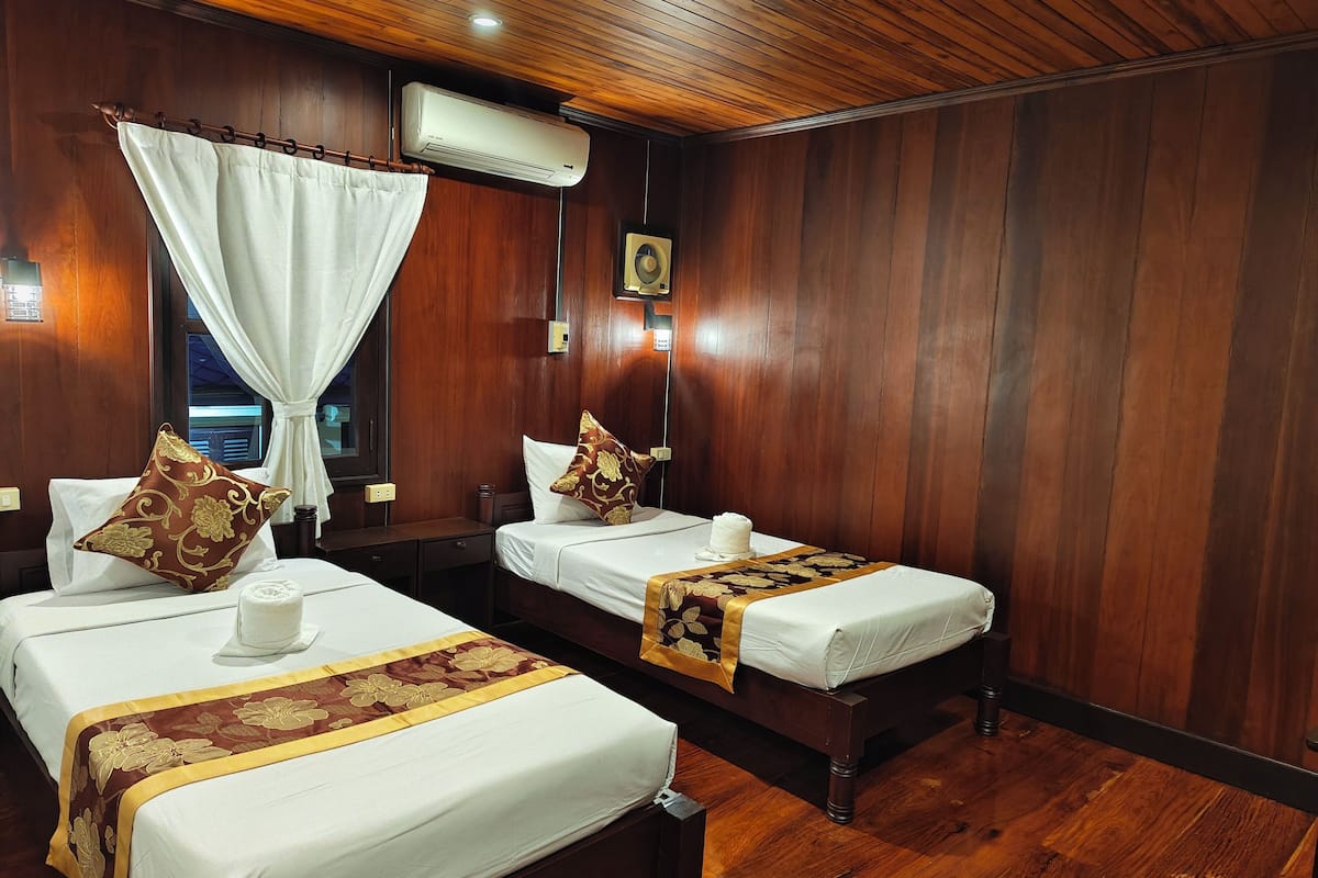 A hotel room with two beds, wooden flooring, a ceiling fan, and an air conditioning unit.