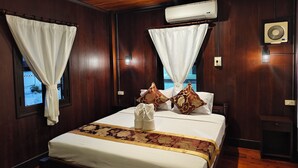 A hotel room with a wooden ceiling, a bed with white bedding and floral-patterned pillows, an air conditioning unit, and a window with a curtain.