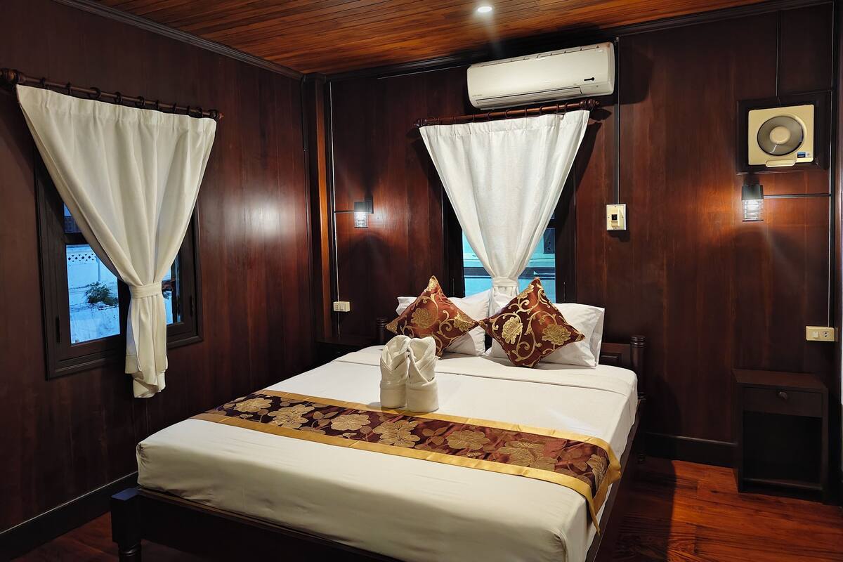 A hotel room with a wooden ceiling, a bed with white bedding and floral-patterned pillows, an air conditioning unit, and a window with a curtain.