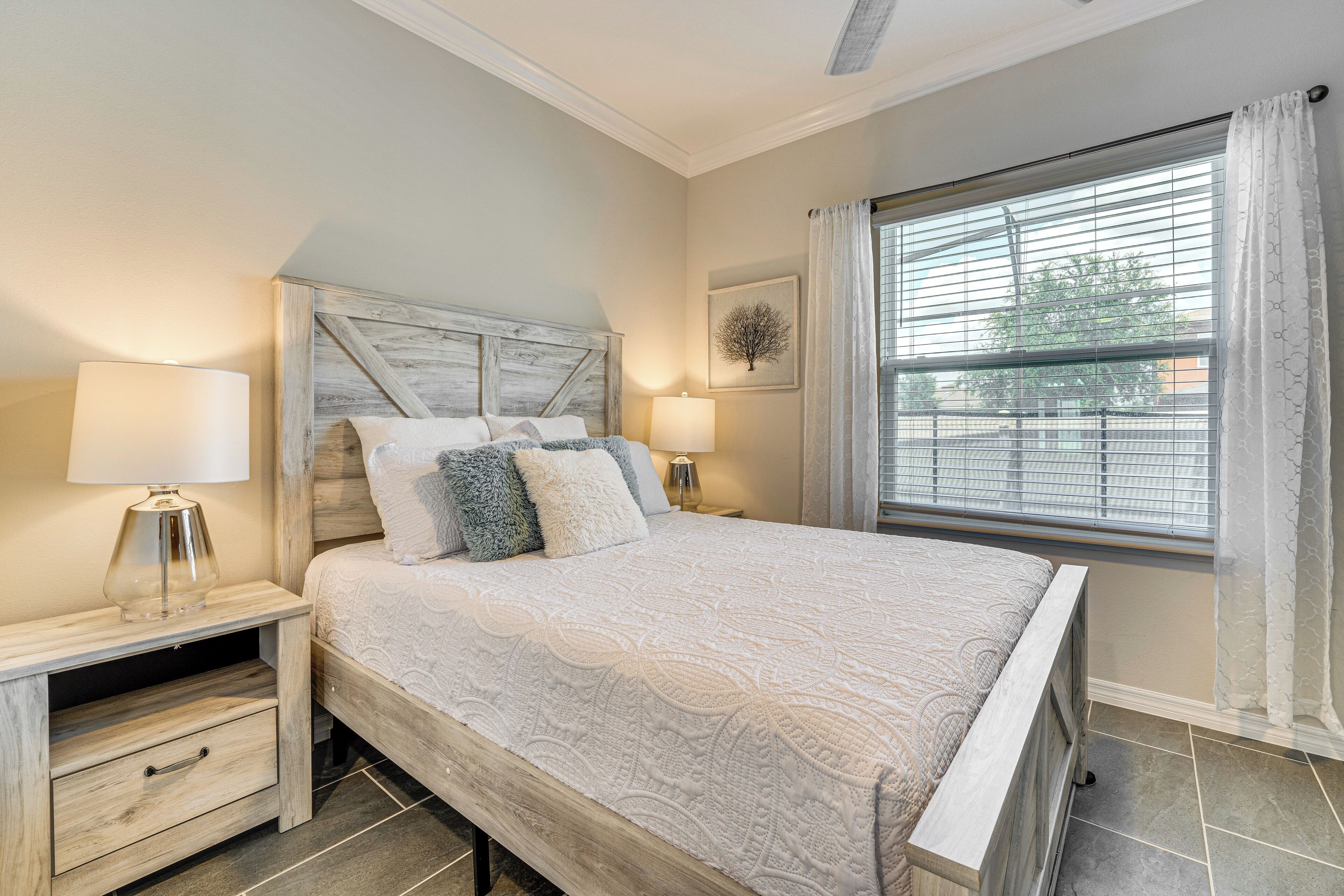 A bedroom with a large bed, two bedside tables, a nightstand, a window with blinds, and a wall-mounted lamp.