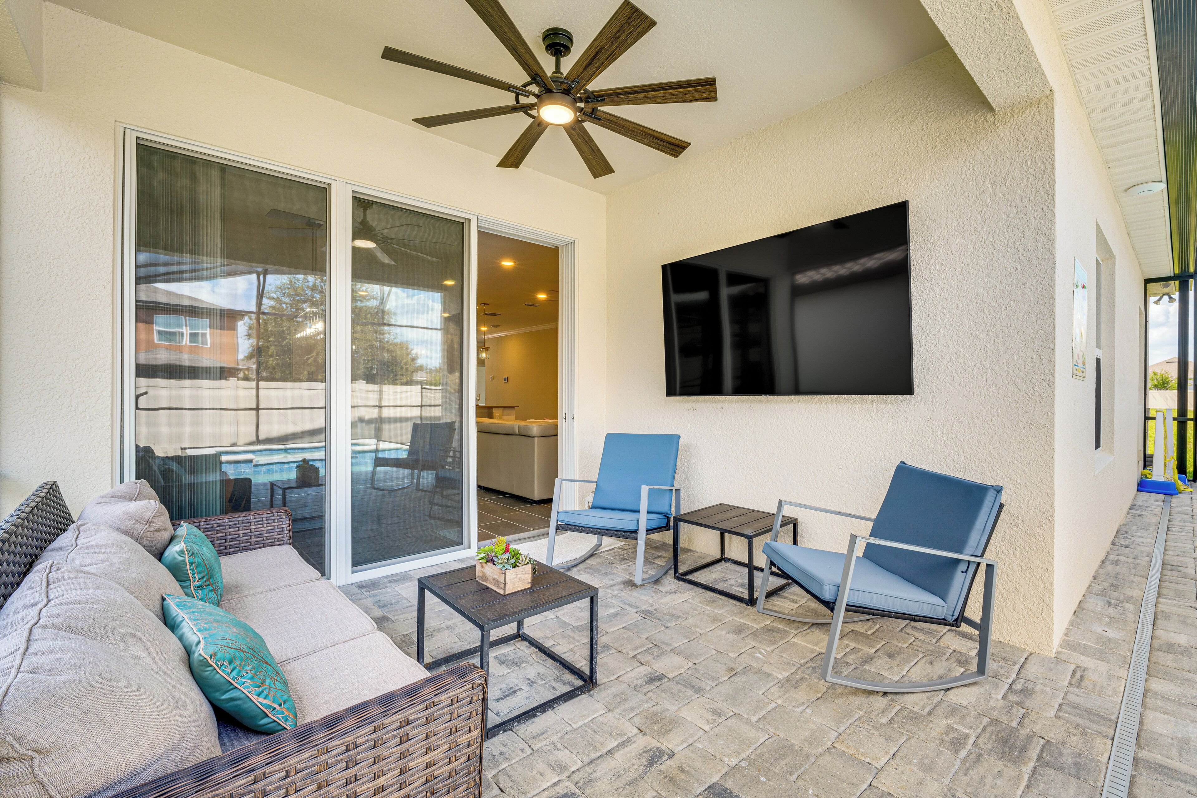 Un patio couvert doté d’un ventilateur de plafond, d’un canapé avec des coussins, de deux fauteuils, d’une petite table avec une plante et d’un téléviseur fixé au mur.