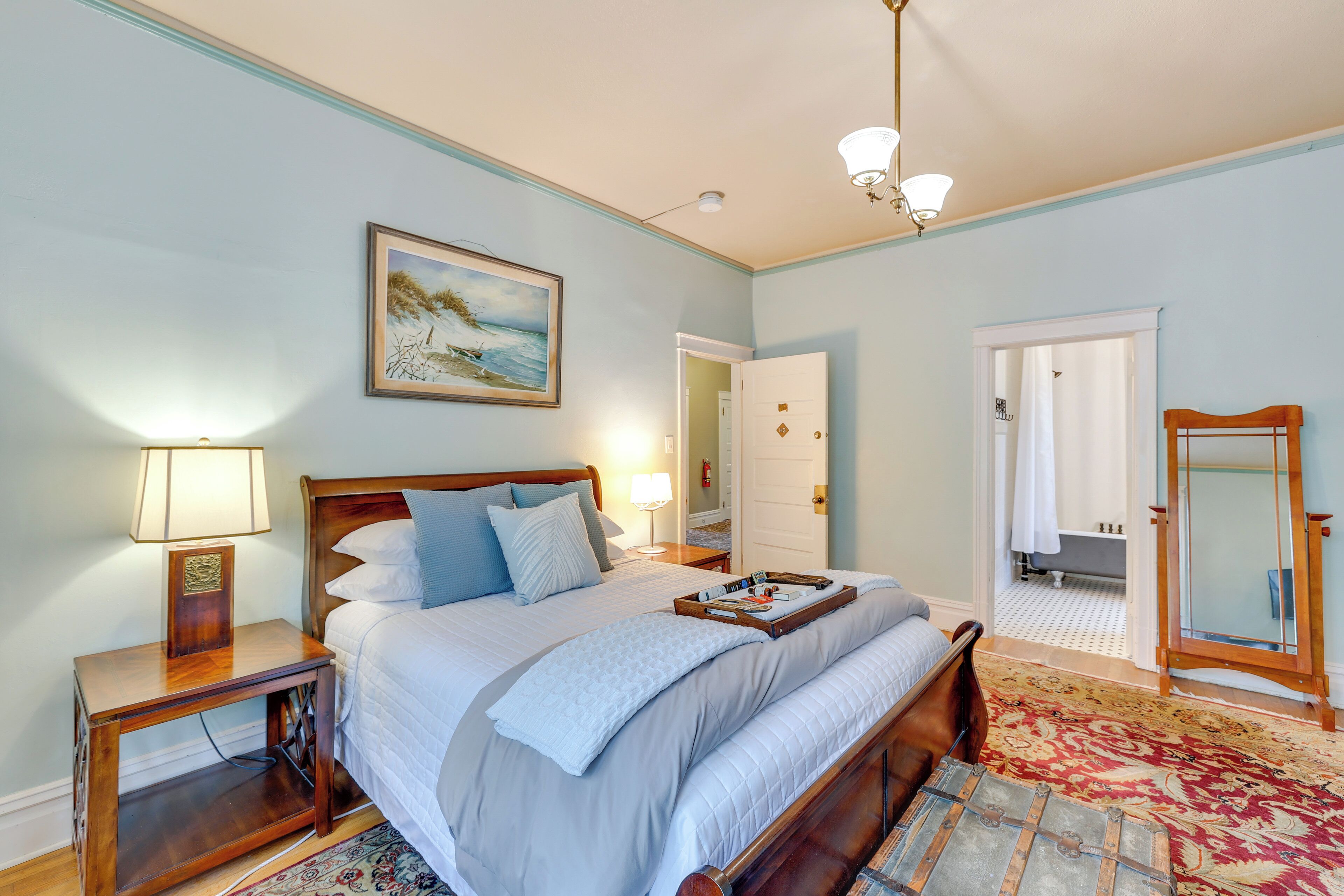 A bedroom with a bed, bedside tables, a nightstand, a lamp, a painting, a mirror, and a bathroom door.