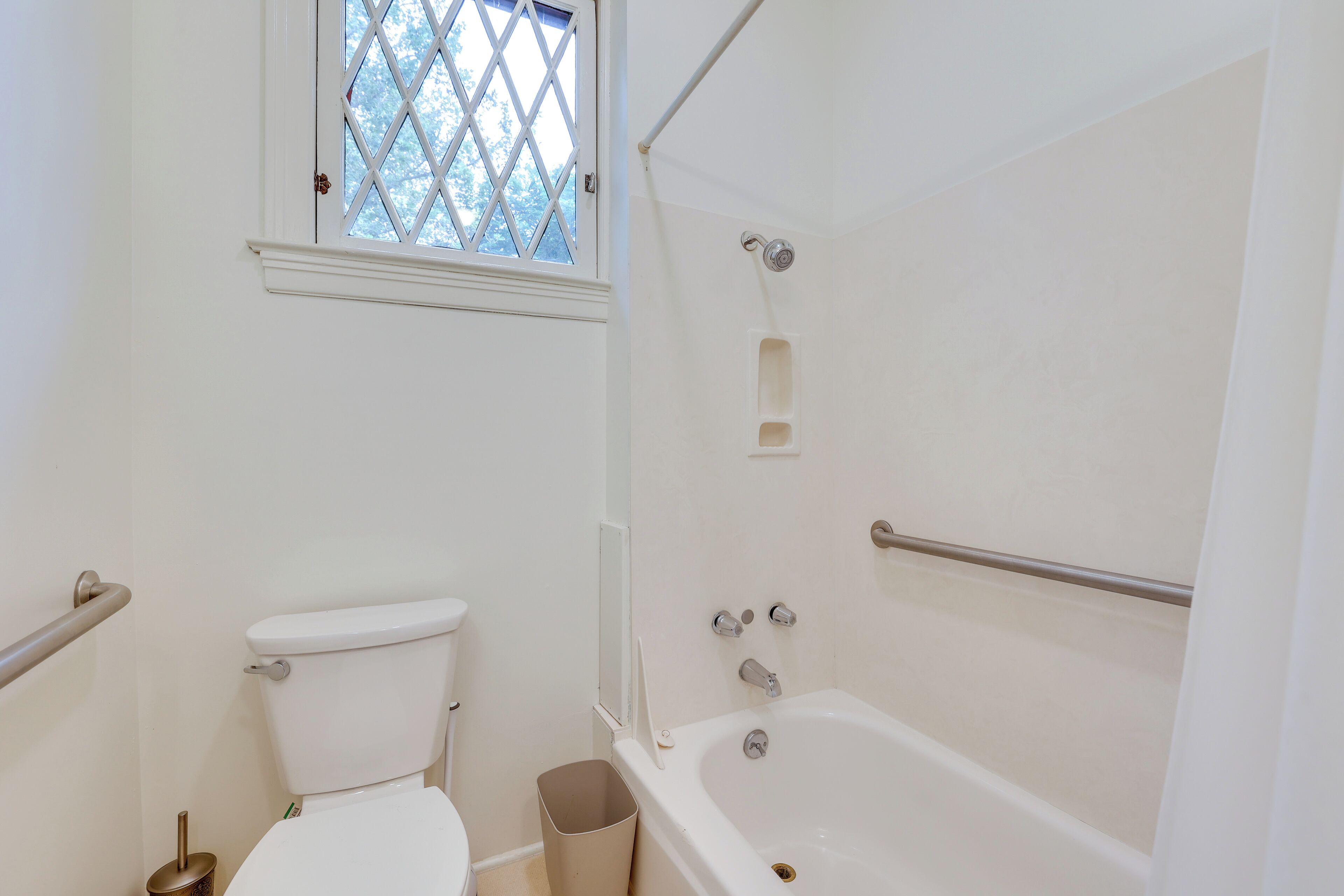 A bathroom with a toilet, a bathtub, a window, and a trash bin.
