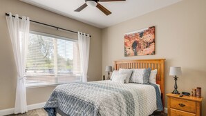 Une chambre à coucher avec un lit, une table de chevet, un ventilateur de plafond, une fenêtre avec des rideaux et un tableau au mur.