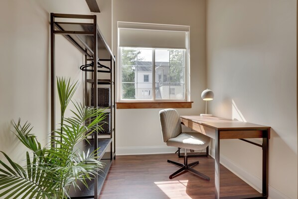 A modern room with a wooden desk, a chair, a window with a view of buildings, and a plant on the left. - Modern 3BR Home Near JMU - EMU Sleeps 6 (Harrisonburg)