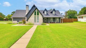 A two-story house with a front lawn, a pathway leading to the entrance, and a wooden fence in the background. - Fire Pit + Sauna: Spacious Lake Texoma Retreat! (Denison)