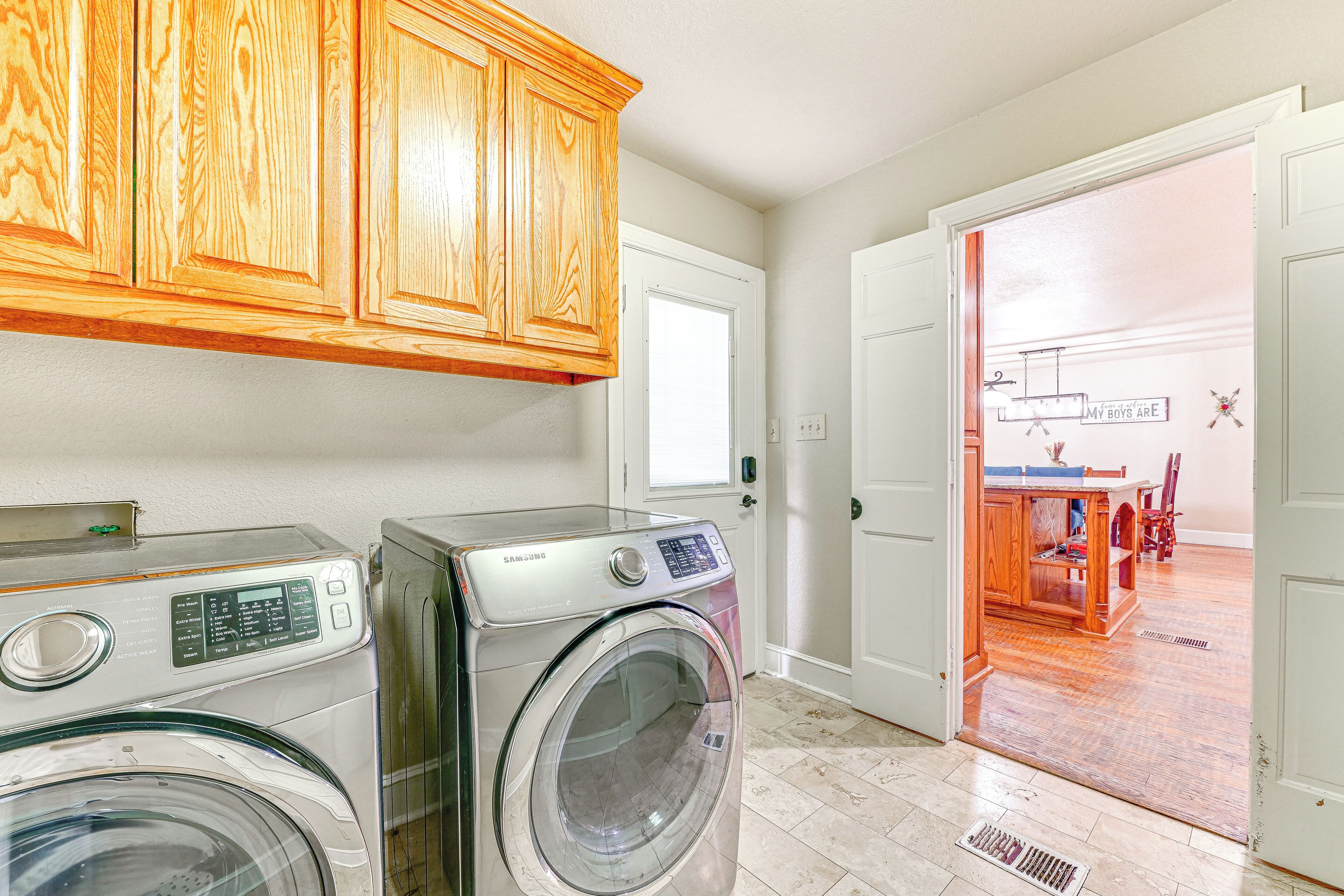 A laundry area with two front-loading washing machines, wooden cabinets, and a view into a dining area through an open door.