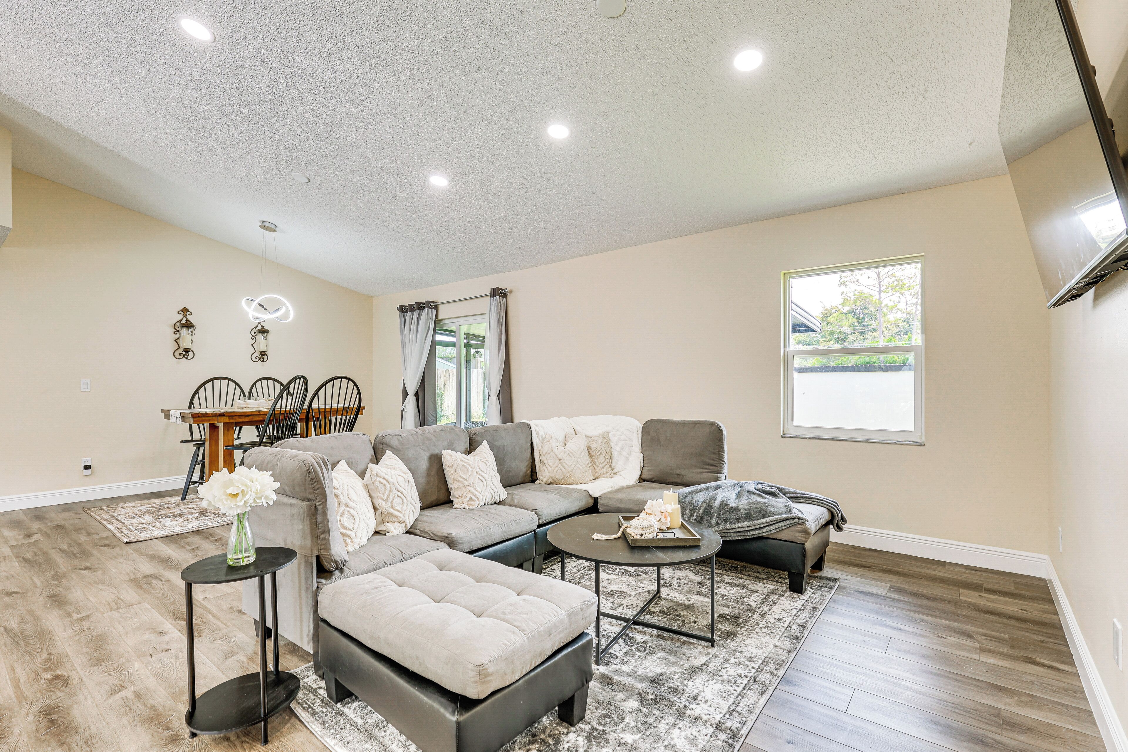 A spacious living room with a sectional sofa, ottoman, and coffee table. There is a dining area with a table and chairs, and a window providing natural light.
