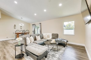A spacious living room with a sectional sofa, ottoman, and coffee table. There is a dining area with a table and chairs, and a window providing natural light. - 16 Mi to Downtown Tampa: Home w/ Hot Tub! (Brandon)