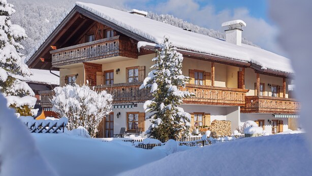A multi-story chalet with wooden balconies, snow-covered surroundings, and a mountain backdrop. - Cozy 1-room apartment with terrace and mountain view (Oberau)