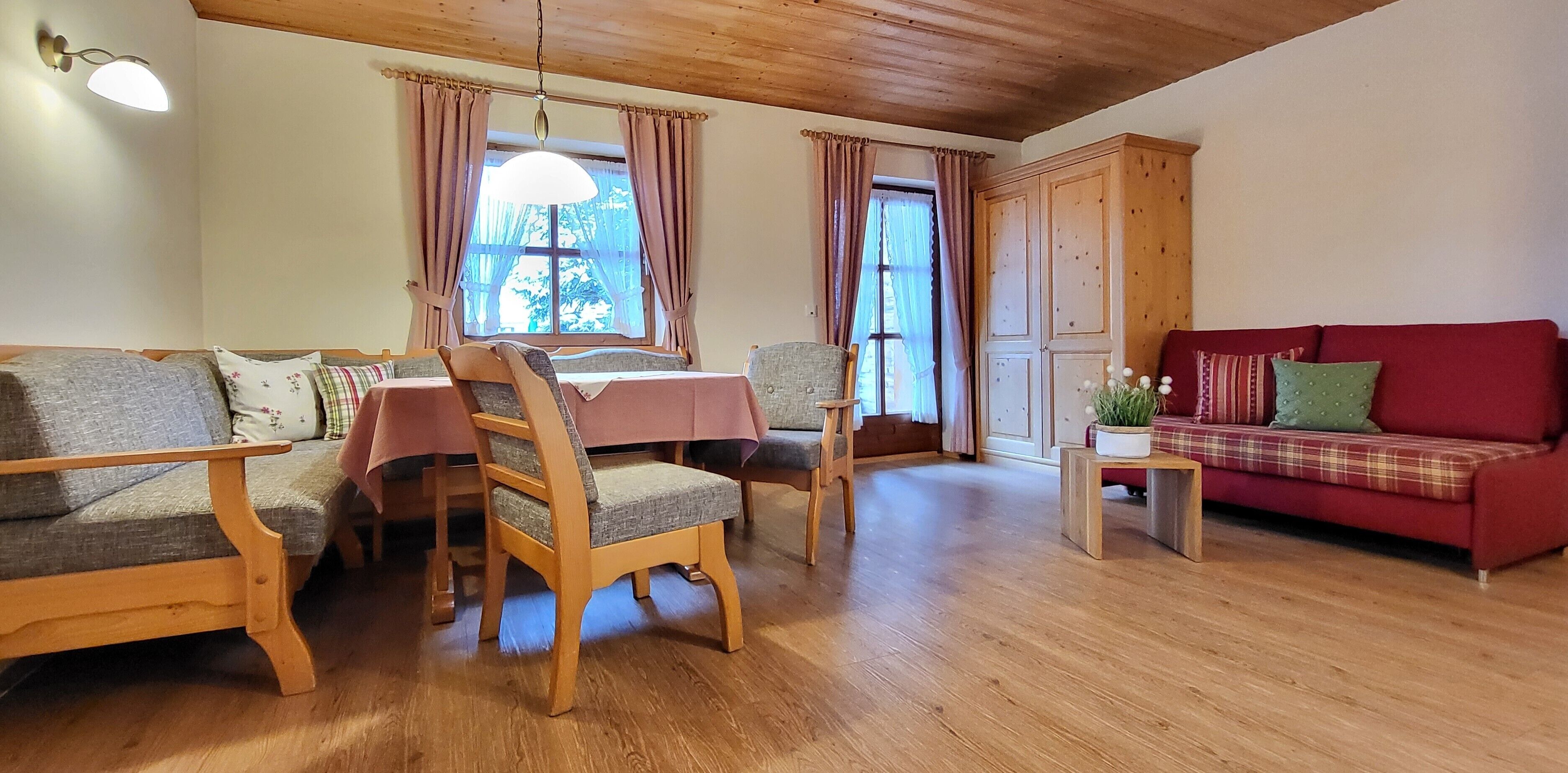 A cozy living room with wooden furniture, a red sofa, a dining table with chairs, and a wooden cabinet.