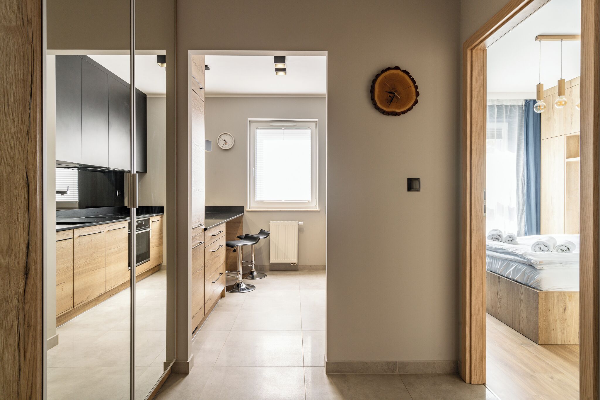 Une cuisine moderne avec des armoires en bois, un comptoir noir et une fenêtre avec des stores. Il y a une horloge au mur et une porte en bois qui donne sur une chambre à coucher, où se trouvent un lit et une fenêtre.